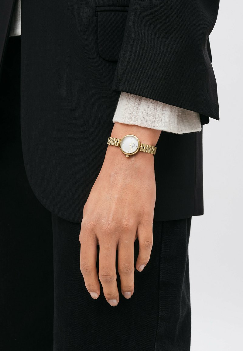 Montre dorée avec un cadran rond blanc, bracelet à maillons et design minimaliste. Portée au poignet, assortie à un blazer noir et un pull léger.
