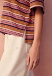 Gestreiftes Kurzarmshirt in Violett- und Orangetönen mit Seitenschlitzen, kombiniert mit beigen High-Waist-Hosen und einem goldenen Ring an der Hand.