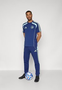 Marineblå idrettstrøye og bukser med lyseblå detaljer, Adidas-logoer og et sirkulært emblem; står ved siden av en flerfarget fotball.