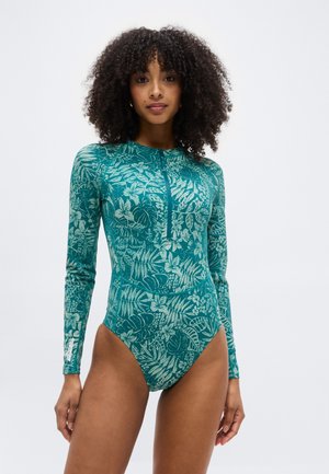 Γυναίκα με σγουρά μαλλιά, ντυμένη με ένα μπλε-πράσινο κορμάκι με μακριά μανίκια και ανθικό μοτίβο, με φερμουάρ στην πρόσοψη, στέκεται μπροστά σε ένα απλό λευκό φόντο.