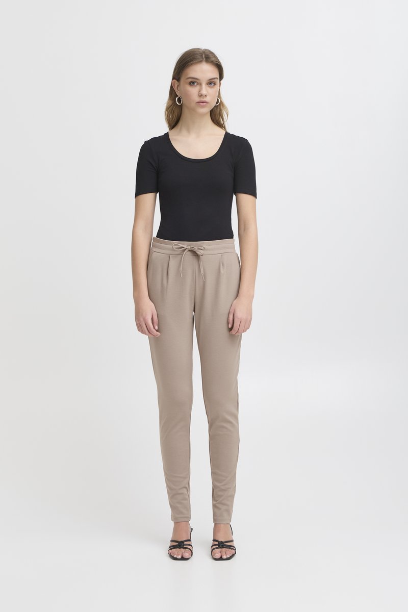 Giovane donna con top nero a maniche corte, pantaloni beige con coulisse, orecchini a cerchio e sandali neri con cinturini, in piedi davanti a uno sfondo bianco uniforme.