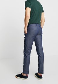 Tmavě modré kalhoty na míru se slim fit střih, spárované se zelenou košili s krátkým rukávem a černými teniskami. Zadní pohled ukazuje úhledné švy a kapsy.