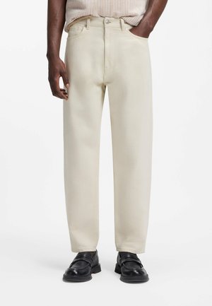 Mand iført beige straight-leg jeans, sorte læder loafers og en lys strik top, hænderne i lommerne, stående på hvid baggrund.