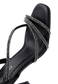 Schwarze Sandale mit quadratischer Zehenpartie und einem eleganten, strukturierten Fußbett. Mit mehreren überkreuzten Riemen, verziert mit Strasssteinen für ein glitzerndes Detail.
