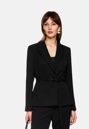 Woman wearing black belted blazer and trousers, with gold circular earrings, holding a black handbag against a plain light background.