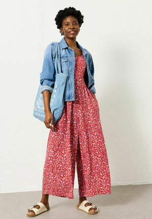 Woman wearing a red floral maxi dress and denim jacket, carrying a blue patterned tote bag, standing on a gray floor against a white wall.