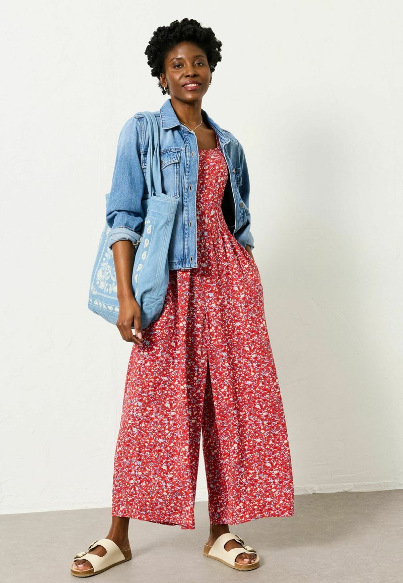 Ženska v rdeči maxi obleki z rožnatim vzorcem in jeans jakni, ki nosi modro vzorčasto torbo, stoji na sivi tleh ob beli steni.