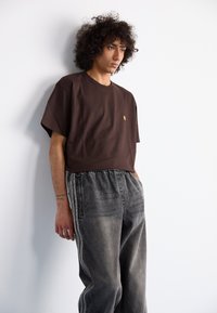 Jonge man met krullend haar, gekleed in een bruine t-shirt en losse grijze jeans, staat tegen een witte muur met zijn handen in zijn zakken.