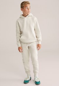Felpa color beige chiaro con tasca frontale e pantaloni jogger coordinati. Entrambi realizzati in materiale morbido. Le sneakers presentano dettagli in teal e bianco.