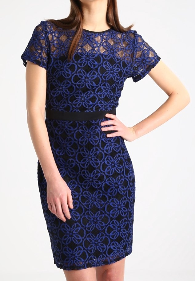 Žena v modrých krajkových šatech po kolena s krátkým rukávem a květinovými vzory, s černým páskem, stojící s jednou rukou na boku.
