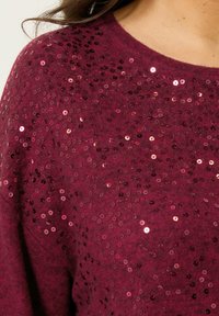 Bordowy sweter o miękkiej fakturze, z rozrzuconymi różowymi i czerwonymi cekinami na przodzie, tworzącymi błyszczący efekt.