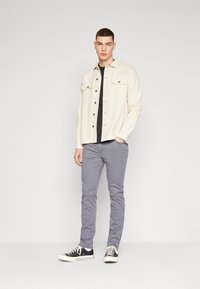 Off-white shirtjacket met twee borstzakken, een zwart T-shirt eronder, grijze slim-fit broek en zwarte sneakers met witte zolen.