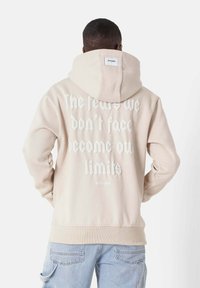 Felpa beige con cappuccio e testo stampato bianco sul retro. Caratterizzata da polsini e orlo a coste, tessuto morbido e una etichetta sul collo.