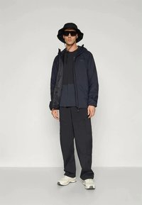 Homem a usar um chapéu tipo balde preto, óculos de sol, casaco preto, calças pretas e ténis brancos, de pé contra um fundo claro e liso.