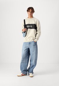 Sudadera beige claro con una franja central negra y un logo blanco, combinada con unos vaqueros azules holgados y unas zapatillas crema con acentos rojos.