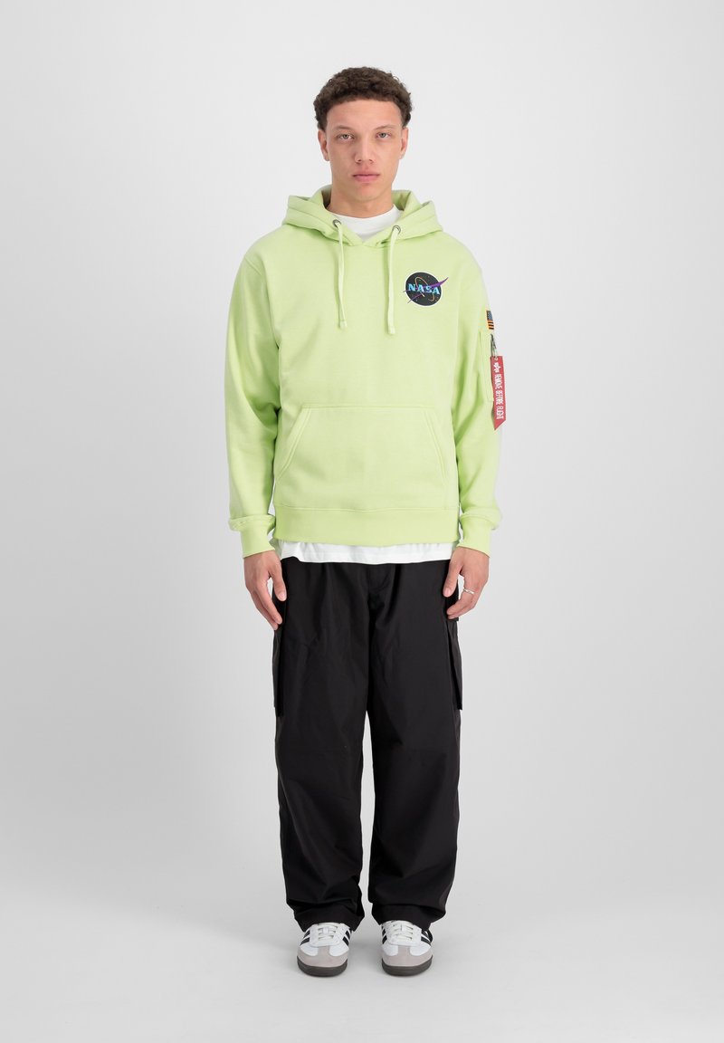 Sudadera verde clara con el logo de NASA, bolsillo tipo canguro y capucha con cordón, combinada con pantalones cargo negros y zapatillas blancas.