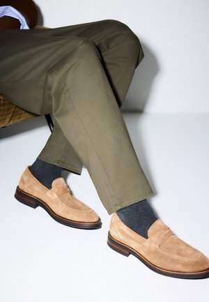 Persona seduta con le gambe incrociate che indossa mocassini in camoscio beige, calze scure e pantaloni verde oliva su sfondo bianco.