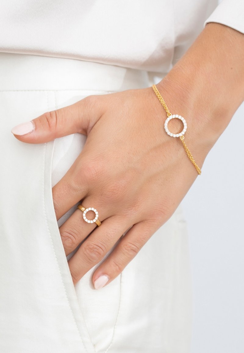 Guldarmband och ring med cirkulära mönster, prydda med klara stenar. Armbandet har en tunn kedja och ringen har ett slätt band.