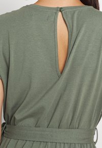 Robe verte à manches courtes avec une ouverture en forme de clé dans le dos, maintenue par un bouton. Fabriquée dans un tissu doux, elle présente une taille ajustée et une jupe plissée.