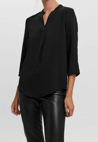 Blouse noire à manches longues avec un col en V, présentant un tissu texturé et un ourlet légèrement incurvé. Associée à un pantalon en cuir noir brillant.
