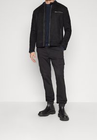 Giacca in camoscio nero con zip frontale, maglione in maglia textured blu navy, pantaloni cargo neri e stivali chunky neri. Outfit casual con dettagli funzionali.