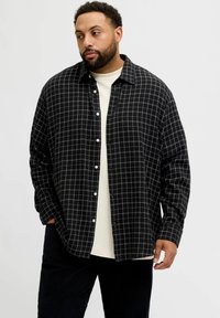 Mees habemega, kes kannab must-valget ruudulist nööbiga särki valge T-särgi peal, seisab ühel käel taskus, seistes lihtsa tausta ees.