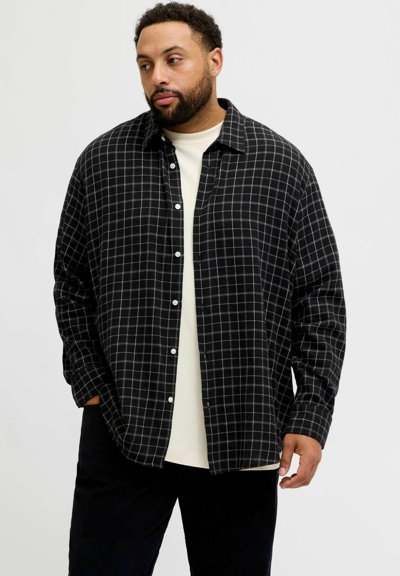 Mees habemega, kes kannab must-valget ruudulist nööbiga särki valge T-särgi peal, seisab ühel käel taskus, seistes lihtsa tausta ees.