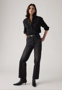 Zwarte spijkerblouse, zwarte high-waisted jeans en zwarte enkellaarzen. Soepele textuur, getailleerd ontwerp en taps toelopende pijpen met een subtiele glans.