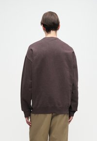 Hanorac din fleece maroniu cu o croială lejeră, de tip crew neck și umeri căzuți. Include manșete și tiv ribbate, purtat cu pantaloni khaki.