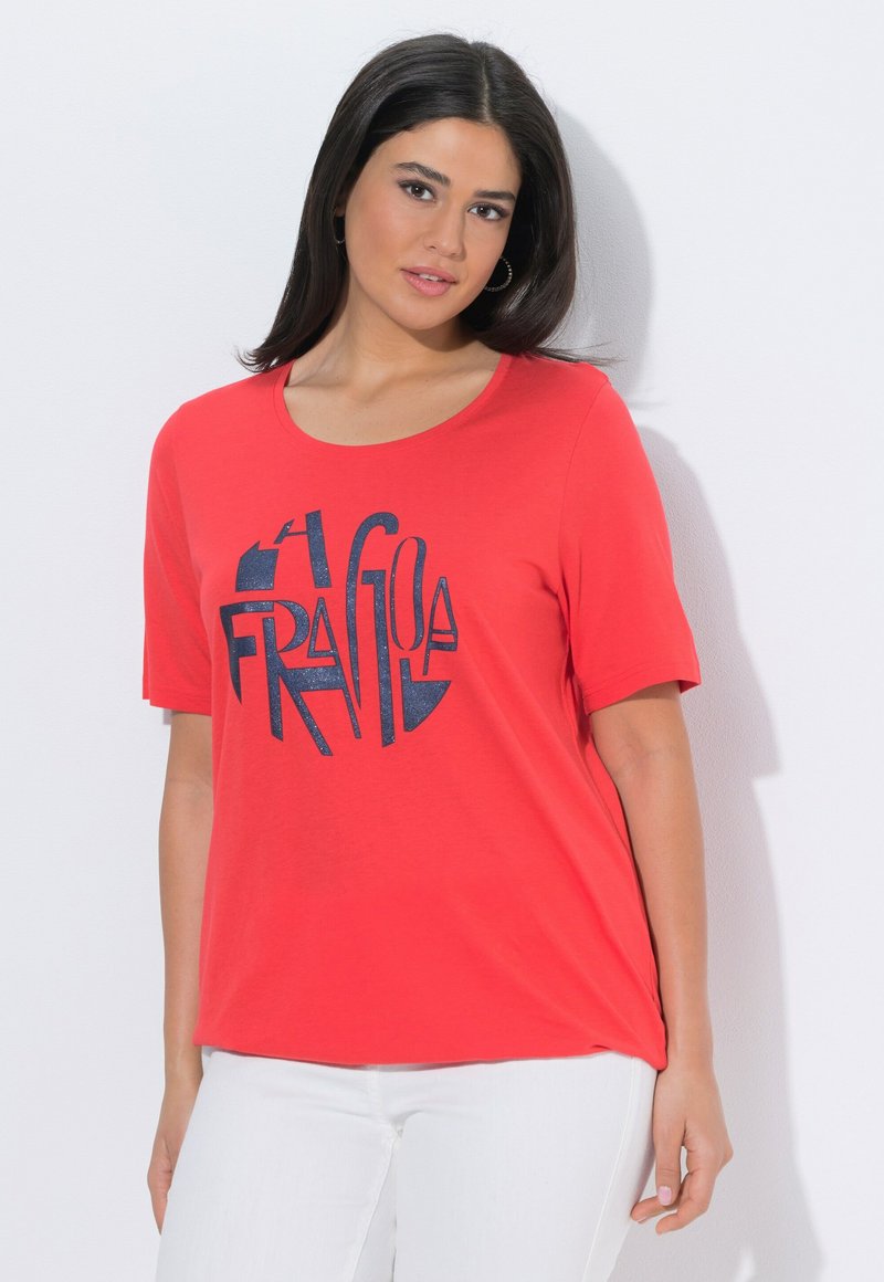 Mujer con cabello liso y oscuro, vistiendo una camiseta roja con texto abstracto azul oscuro y pantalones blancos, posando frente a un fondo blanco.