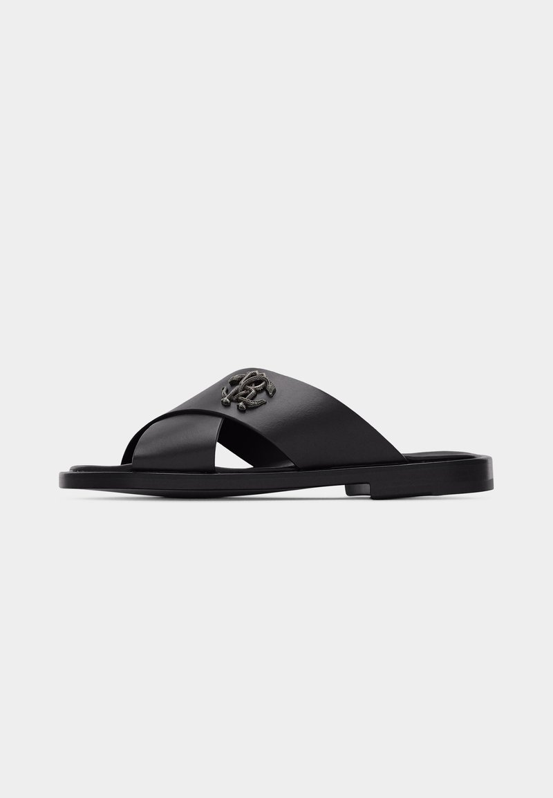 Μαύρο δερμάτινο σανδάλι τύπου slide με διασταυρωμένα λουριά και ένα μικρό μεταλλικό διακοσμητικό έμβλημα στο πάνω λουρί, με επίπεδη σόλα.