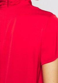 Blouse en soie rouge avec un col montant et des manches courtes. Présente une texture lisse et une brillance subtile, s'arrêtant à l'épaule.