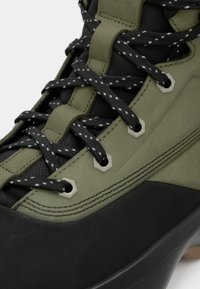 Πράσινα και μαύρα ορειβατικά παπούτσια με υφή στην άνω επιφάνεια, ενισχυμένο σύστημα κορδονιών και καουτσούκ σόλα. Τα ασημένια αξεσουάρ προσθέτουν αντοχή.