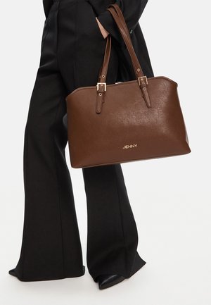 Osoba oblečená v černých širokých kalhotách a černých botách držící velkou hnědou koženou tašku přes rameno se dvěma uchy a zlatým kováním.