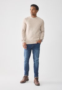 Beige gepunkteter Strickpullover, schmale blaue Jeans und braune Lederstiefel. Das Model steht mit den Händen in den Taschen vor einem hellen Hintergrund.