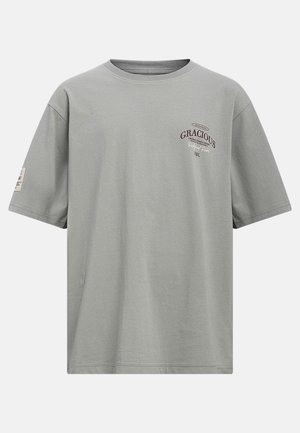 T-shirt gris manches courtes col rond avec le texte "GRACIOUS", petit logo sur la poitrine gauche et un petit patch sur la manche gauche.