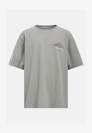 T-shirt gris manches courtes col rond avec le texte "GRACIOUS", petit logo sur la poitrine gauche et un petit patch sur la manche gauche.