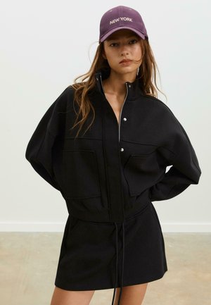 Zwarte zip-up jas met oversized mouwen, voorzien van twee voorzakken en een trekkoord in de taille, gecombineerd met een paarse "NEW YORK" pet.
