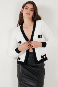 Žena koja namješta bijeli kardigan s crnom obrubom i gumbima, nosi crnu kožnu suknju, stoji uz jednobojni bijeli zid.