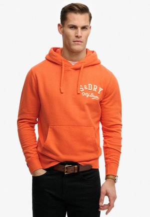Mand iført orange hættetrøje med teksten "S&DRY NY State", sorte bukser, brunt bælte og ur, stående med den ene hånd i lommen.
