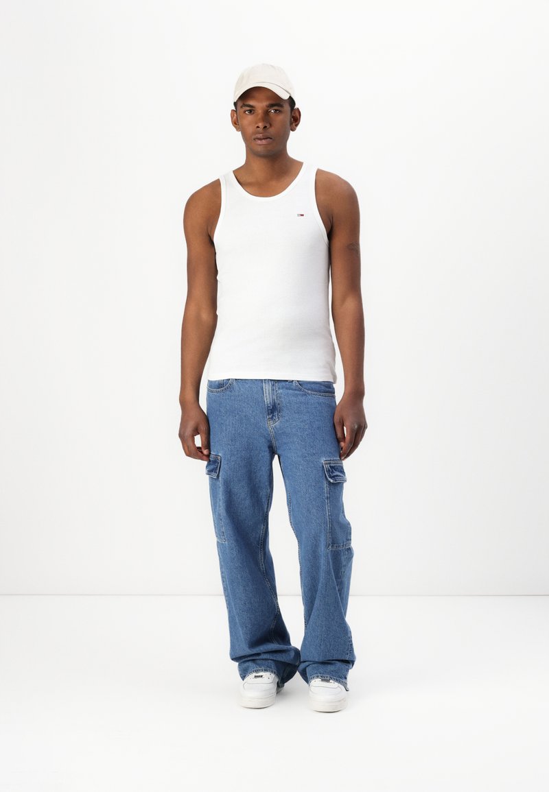 Hvit tanktopp med lite logo, blå denim cargo bukser med sidelommer, og hvite joggesko toppet med en lyse farget caps.