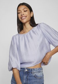 Bluză scurtă de culoare lavandă deschisă, cu mâneci bufante, confecționată dintr-un material texturat, asortată cu blugi din denim cu talie înaltă și un nasture simplu la talie.
