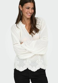 Usmívající se žena s dlouhými hnědými vlasy, oblečená v bílé průhledné blůze s květinovou výšivkou a černými kalhotami, s rukama volně zkříženýma.