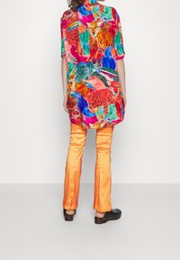 Person som bär en färgglad skjorta med djurmönster, korta ärmar, glänsande orange byxor och svarta klackskor, stående mot en enkel vit bakgrund.