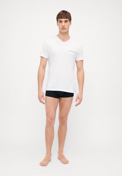 Tricou alb cu decolteu în V și un mic logo negru pe pieptul stâng, asortat cu chiloți negri; modelul stă desculț pe un fundal simplu.