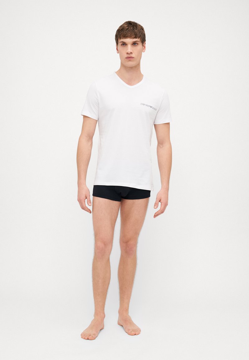 White V-neck T-shirt with a small black logo on the left chest, paired with black briefs; the model stands barefoot against a plain background.