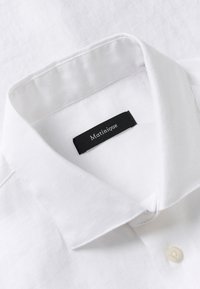 Ovratnik bele lanene srajce s črno etiketo, na kateri je zapisano ime znamke "Matinique", ter vidnim gumbom pod ovratnikom.