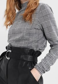 Svartvit rutiga långärmad topp med hög krage, parat med högmidjade svarta byxor i fuskläder som har ett brett bälte och silverfästen.