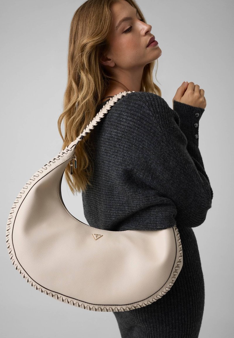 Femme portant un pull en maille foncée porte un grand sac à épaule crème en forme de croissant avec des bords cousus et un petit logo triangulaire.