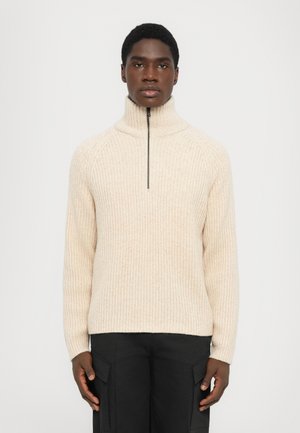 Light beige ribbed sweater with a high collar and half-zip closure, made from a soft fabric. Paired with black cargo pants.